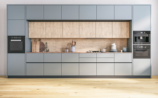A front view of a modern luxury kitchen with high grayish / blue and hardwood kitchen cabinets with an empty space in front, on the oak parquet hardwood floor. Kitchen utensils, a faucet, a sink, a bulit in oven and coffee maker, a large pile of plates, cutting boards, a pan on the stove in front of hardwood  backsplash. 3D rendered image.