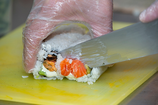 A gloved hand cuts sushi on a cutting board.