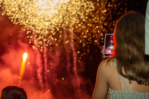 Castilla La Mancha, Spain. Rear view of a young woman photographing fireworks with her mobile phone during the celebration of popular festivities in honor of the town's patron saint.
