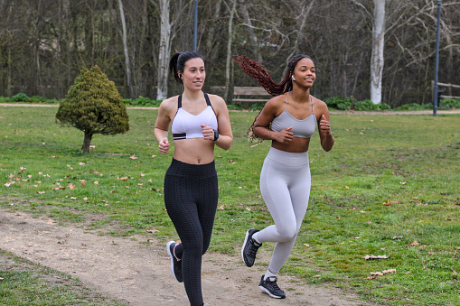 Young caucasian woman and young black woman running together outdoors in sports clothes, smiling, multiethnic, front view, with nature in background. Concept of sport in company