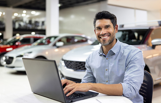 Happy Latin American salesman working on a laptop at a car dealership and looking at the camera smiling