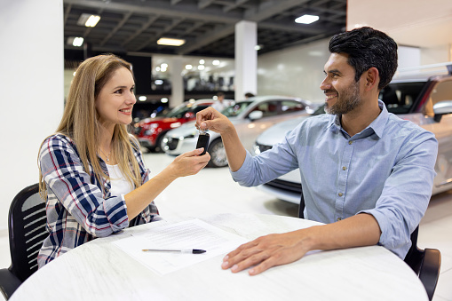 Happy Latin American woman buying a car and receiving the keys at the dealership - car ownership concepts