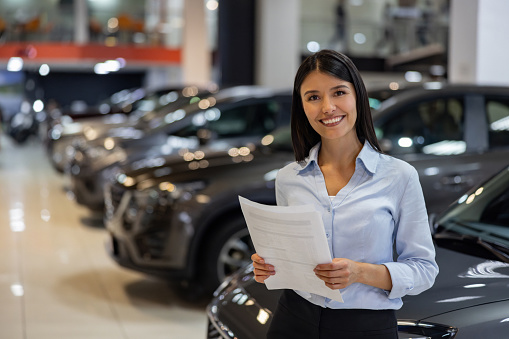 Portrait of a happy car salesperson smiling at a car dealership and looking at the camera holding documents