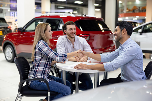 Happy Latin American car salesman handshaking with a couple after closing a deal at the dealership - car ownership concepts