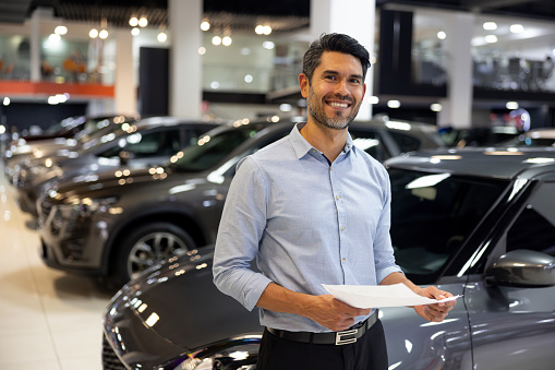 Portrait of a happy Latin American salesman working at a car dealership holding a lease agreement and looking at the camera smiling