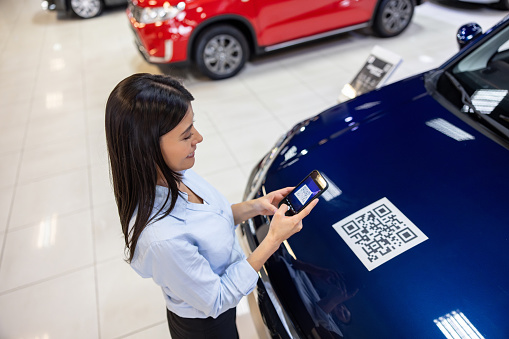 Happy Latin American car salesperson scanning a QR code on a car at the dealership using a mobile phone