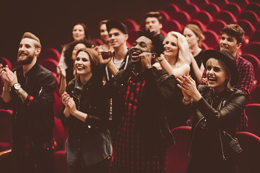 Multiracial group of excited young people in the cinema or theater, watching, laughing, clapping hands.