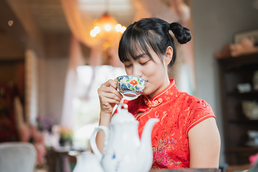 Experience the fusion of fashion and tradition as a young Asian woman, adorned in Chinese dress, delicately prepares and enjoys hot tea.