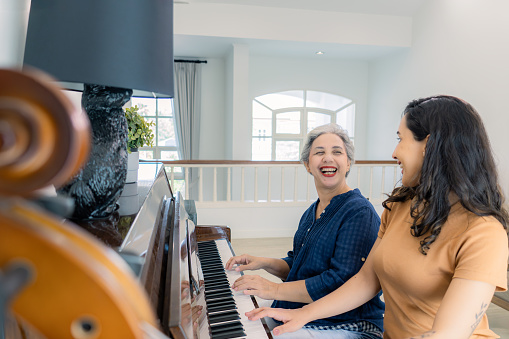 A mixed-race mid-adult daughter joyfully playing piano with her healthy and cheerful Asian senior retired mother on the weekend.