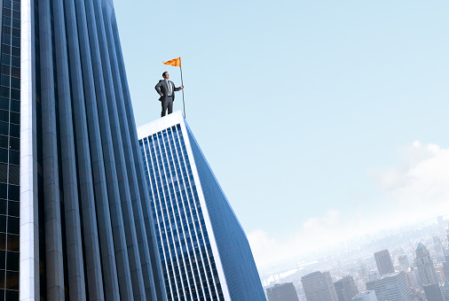 A businessman plants an orange flag on top of a tall skyscraper that oversees the big city below. The blue sky provides ample room for copy or text.