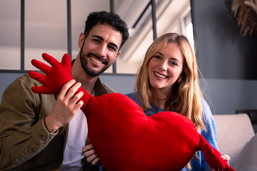 Happy young couple sitting on a sofa holding a heart looking at camera.