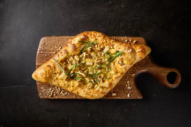Overhead view of Adjarian khachapuri, a modern twist with pear, nuts, and cheese, elegantly displayed on a wooden board against a stark black backdrop.