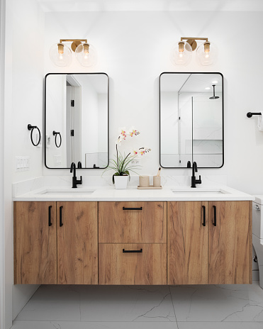 A bathroom with a floating wood vanity cabinet, decorations on the white marble countertop, gold lights mounted above two black mirrors, and black faucets.