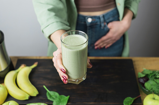 Unrecognizable woman in casual wear holding glass of fresh blended healthy green vegan smoothie. banana, spinach, apple and avocado on a table.