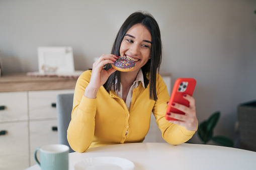 Young female woman having breakfast and using her phone. She is looking cheerful. Eating doughnut. Front view.