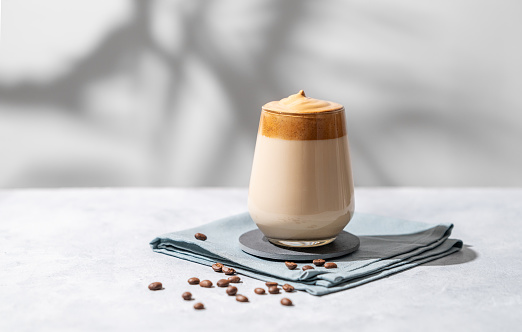 Dalgona coffee. Whipped instant vegan coffee in a glass with beans and shadow on a light background.  The concept of a trendy and popular drink. Free space for text.