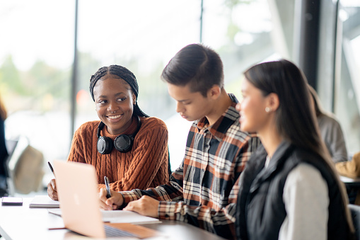 Three University students sit together among their peers as they work together on an assignment.  They are each dressed casually and focused on the task.