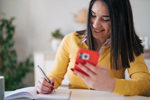 Young female student wearing yellow sweater sitting at the kitchen table and writting down something while using her smart Phone in red case.