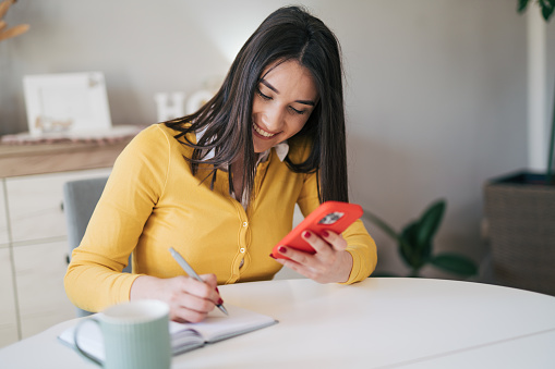 Young female student wearing yellow sweater sitting at the kitchen table and writting down something while using her smart Phone in red case.