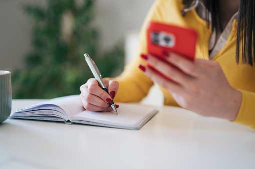 Close up view of young female student wearing yellow sweater sitting at the kitchen table and writting down something while using her smart Phone in red case.