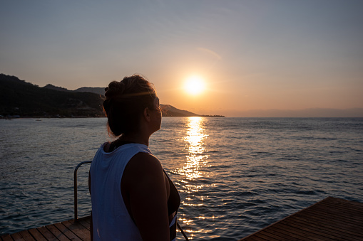 Morning Serenity: Middle-aged woman practicing yoga on a wooden dock at sunrise, greeting the newborn sun on a tranquil summer day.