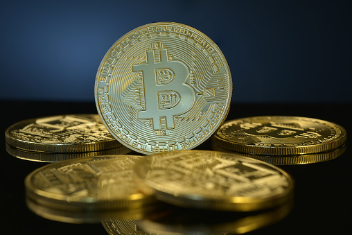 Studio shot of golden Bitcoin isolated on black reflective background, surrounded by pile of coins in soft focus. Bitcoin has a limited supply and  its price is highly volatile. Bitcoin halving event happens every four years.