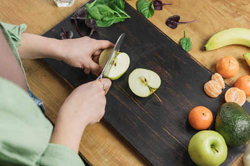 Woman cutting apple, preparing healthy vegan smoothie with spinach leaves, banana and avocado on a table.