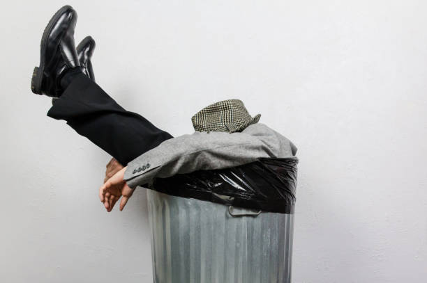 Portrait of Businessman in Suit and Fedora Hat Sitting in Trash Can. Man Thrown Away by Capitalism and Greed.