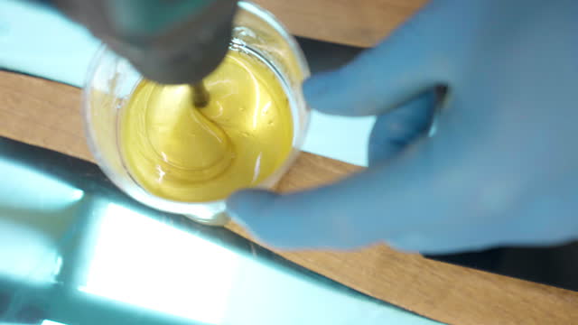 Process of mixing transparent epoxy resin with yellow pigment colour in plastic cup on amazing wooden table with individual design, process of forming unique colour design for wooden furniture and accessories, trendy carpentry artwork