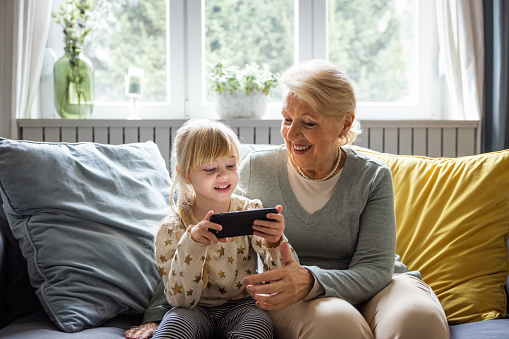 Senior woman sitting with small girl in living room at home using smart phone for video call or playing games.