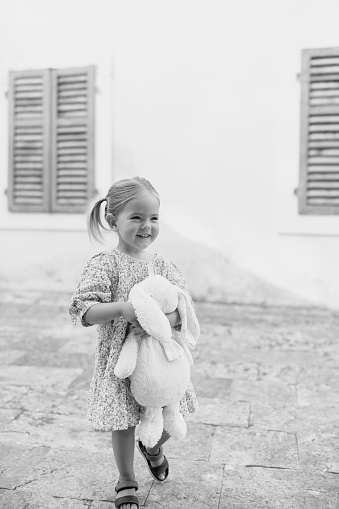 Little smiling girl walks hugging a toy rabbit near the house. Black and white photo. High quality photo