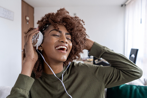 African American Latina woman with afro hair smiling and very happy is sitting on sofa while listening to music with headphones at home. she is dressed in casual clothes. Concept, lifestyle, weekend activity.