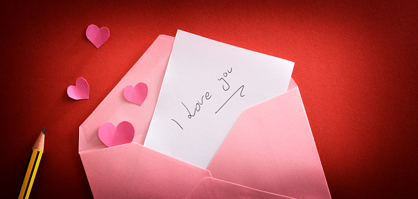 Love note with message I love you in a pink envelope on a red gradient background with pink hearts and pencil. Top view.