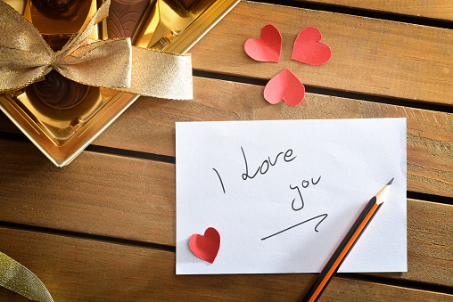 Note I love you written with pencil on wooden table with box of chocolates and red hearts cutouts. Top view.