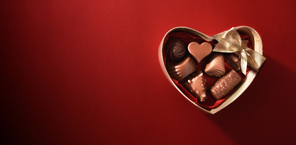 Brown heart-shaped box filled with assorted chocolates with golden bow on red gradient background. Top view.