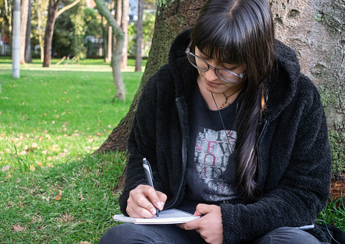 Young girl with glasses sitting writing in nature next to a tree