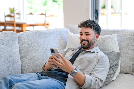 Happy young man relaxing sitting and reclining on the sofa at home. He is using a mobile phone.