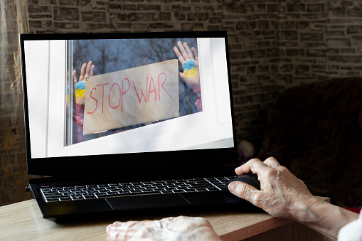 A girl in the window of the house holds a poster Stop the war and a painted flag of Ukraine in her hands, as seen in the photo from a laptop at home