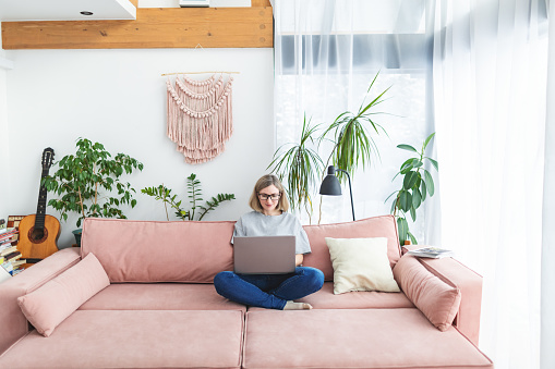 Mid adult woman working from home using laptop sitting on sofa at living room