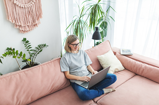 Mid adult woman working from home using laptop sitting on sofa at living room