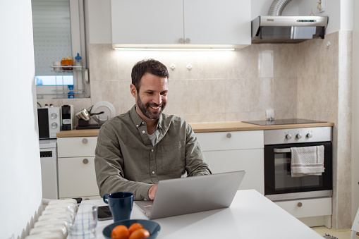 Photo of a handsome man working at home. He is sitting at the dining table in the kitchen and typing on the laptop keyboard.