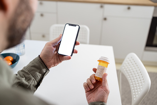 The man's hands hold a pill bottle and a smart phone with a white blank screen. Minimalistic composition with copy space. Nutrition recommendations. Healthy technology. Capsule medicine. Telemedicine. Assistance.