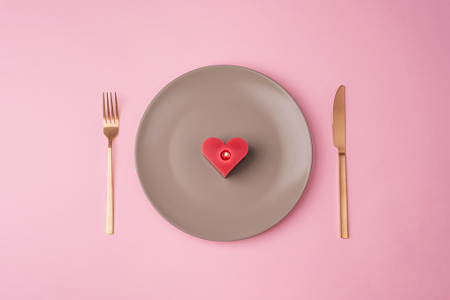 Gray plate with red candle in a shape of heart, fork and knife on pink background. Valentines day concept. Top view, flat lay.