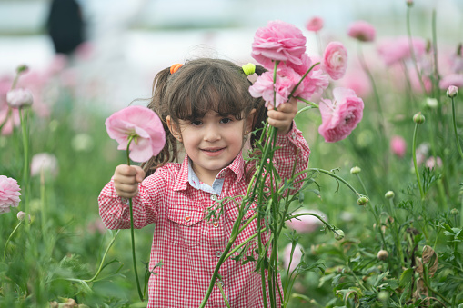 Cute little kid girl 3-4 year old with long curly red hair wear floral wreath and stylish rustic dress over nature background in garden outdoor. Springtime. Smiling baby with flowers. Childhood.