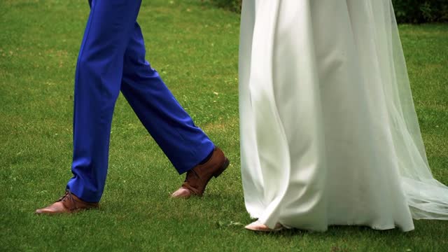 Closeup shot of the legs of bride and groom walking together on the grass in the park on the sunny wedding day, slow motion