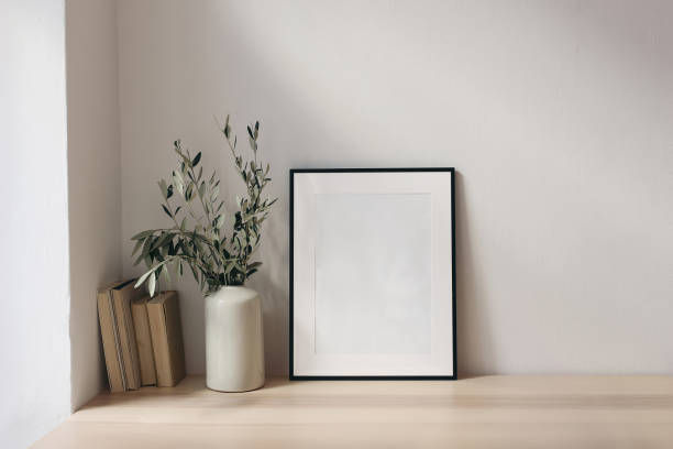 istock Elegant Mediterranean working space. Books, empty vertical black picture frame mockup. White wall background. Wooden desk, table. Vase with olive branches. Home office concept. Scandinavian interior design. 1978513142