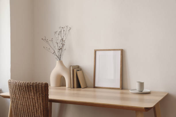 istock Empty wooden picture frame mockup on wooden table. Beige wall background in sunlight. Boho shaped vase, dry flowers on table. Cup of coffee, old books. Working space, home office. Art, poster display. 1978514180