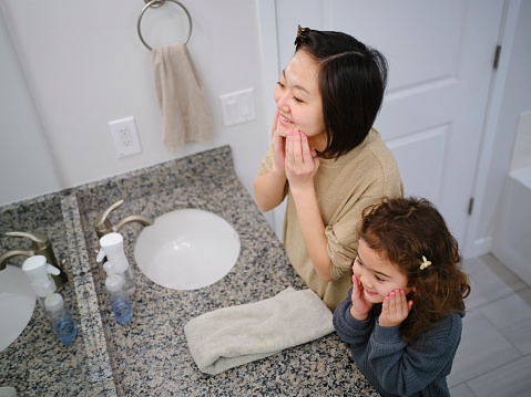 A Japanese mother with her multiracial daughter, washing their faces together in a home bathroom.