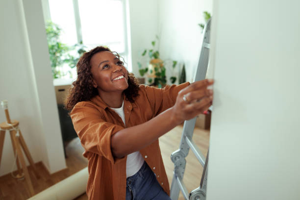 Smiling young woman hanging picture on wall while standing on ladders