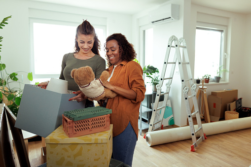 Young lesbian couple unpacking belongings at their new home, one is holding teddy bear toy